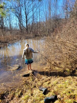 Girl wading in pond with blue tiered skirt, blue boots on shore, blue pond reflecting the sky